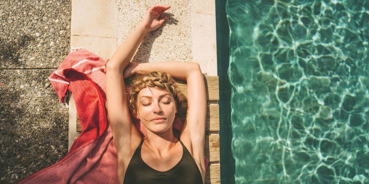 Frau liegt entspannt auf einem Handtuch am Rand eines Pools, lehnt sich mit ausgestreckten Armen zurück und genießt die Sonne neben dem klar schimmernden Wasser