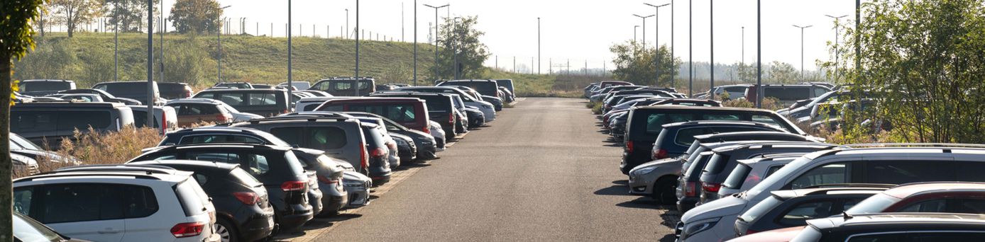 Blick auf einen Parkplatz mit mehreren geparkten Autos auf beiden Seiten eines asphaltierten Wegs, im Hintergrund Bäume und Laternen.