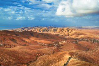 Aussicht vom Mirador de Morro Velosa auf Fuerteventura mit weitläufiger, hügeliger Vulkanlandschaft, serpentinenreicher Straße im Vordergrund und Blick bis zum Atlantik unter bewölktem Himmel