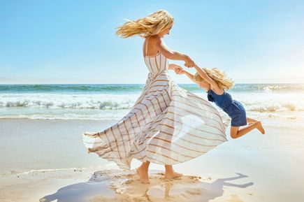 Frau in einem langen, weißen Sommerkleid schwingt ein Kind am Strand