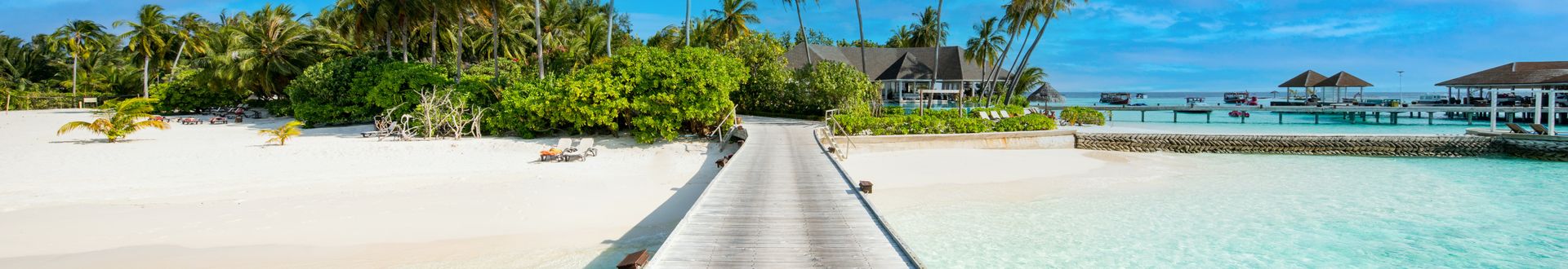 Panoramablick auf einen Holzsteg, der über das Wasser zu einem Resort in den Malediven führt. Umgeben von Palmen und Sandstrand.