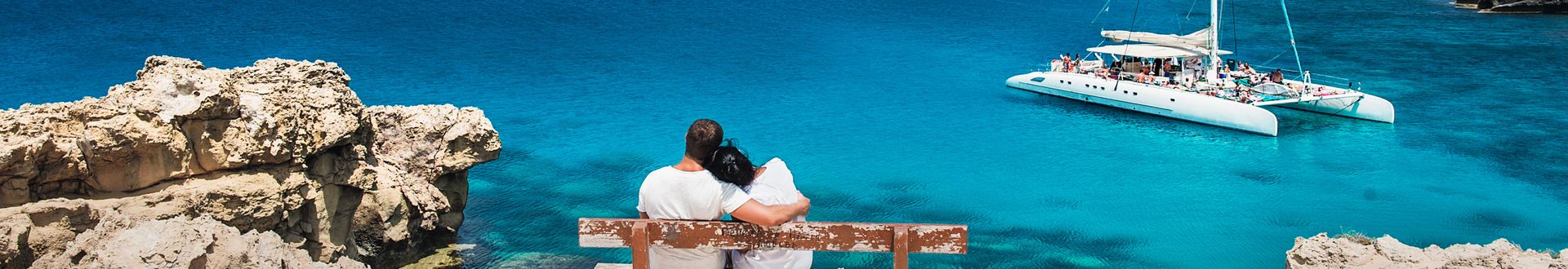 Umarmtes Paar auf einer Bank an der Küste mit Blick über das türkis-blaue Meer und einen Katamaran
