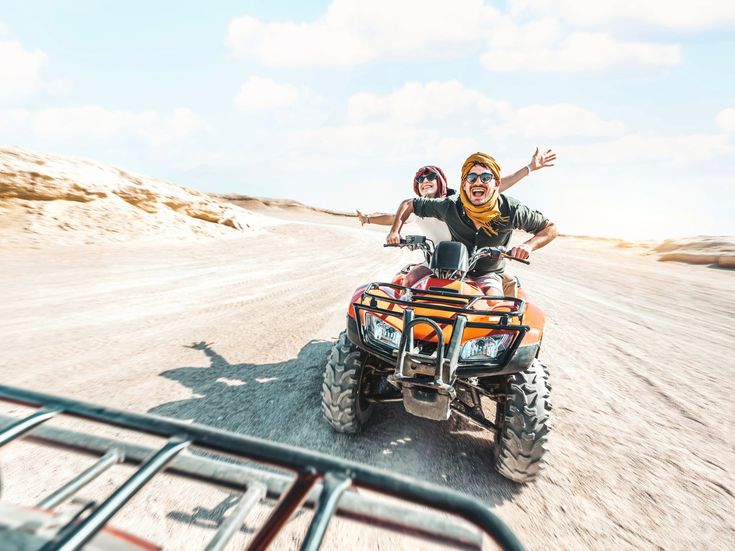 Zwei Menschen fahren lachend auf einem Quad durch eine trockene Wüstenlandschaft