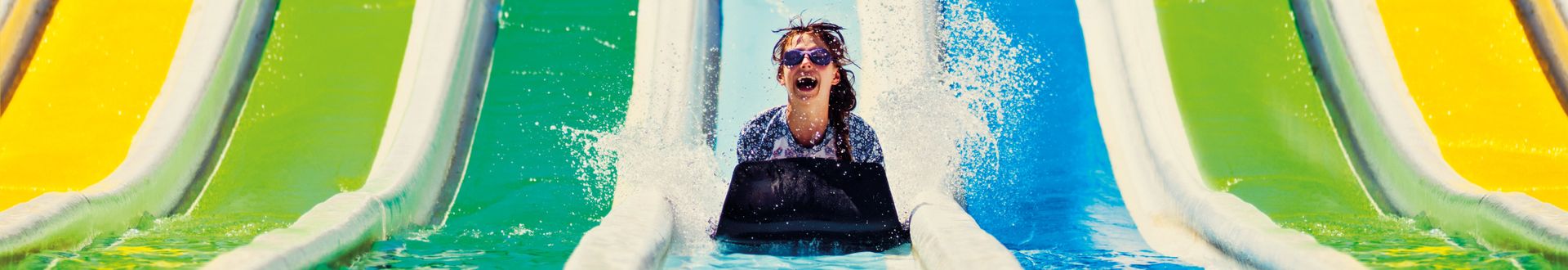Kind rutscht mit einer schwarzen Matte eine bunte Wasserrutsche hinunter und landet spritzend im Pool, umgeben von gelben, grünen und blauen Rutschbahnen