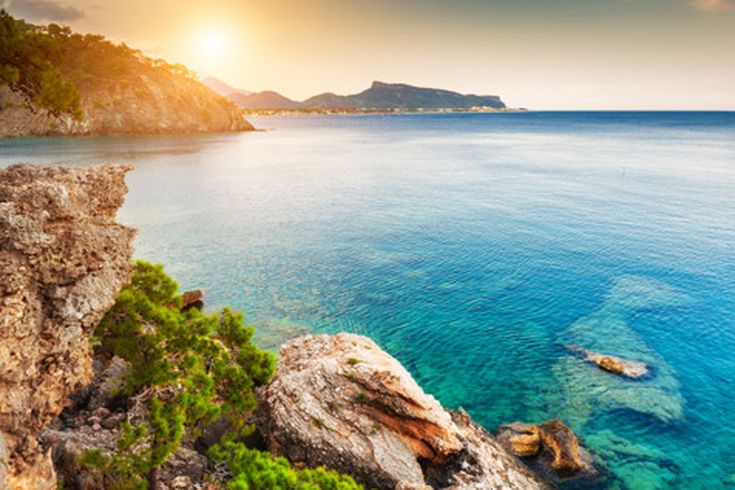 Sonnenuntergang an der Küste von Kemer mit klarem Wasser, Felsen, grünen Pinien und einer ruhigen Bucht
