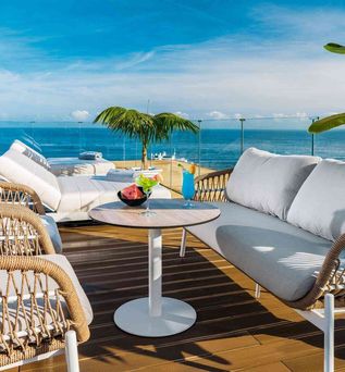 Großer runder Hotelpool mit Liegestühlen und Palmen, angrenzendes Hotelgebäude links, Hügel mit Häusern und Meer im Hintergrund unter blauem Himmel.