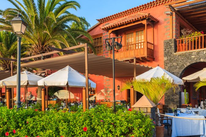 Außenbereich eines Restaurants mit weißen Sonnenschirmen, Tischen mit blauen Tischdecken und einem Gebäude mit roten Wänden und Holzbalkonen im Hintergrund