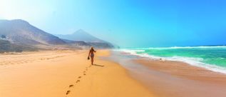 Frau im Bikini mit Sonnenhut läuft am weiten Sandstrand entlang und hinterlässt Fußspuren