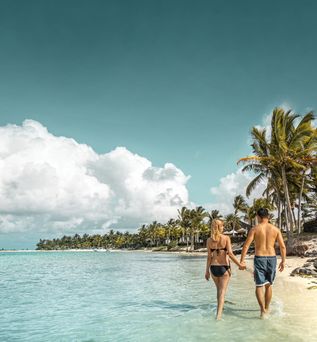 Paar in Badebekleidung hält Händchen und geht am Strand entlang, Palmen und ruhiges Meer im Hintergrund