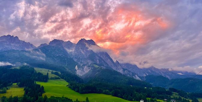 Bergkette im Zillertal mit bewaldeten Hängen und grünen Wiesen unter einem dramatisch orange-violetten Himmel mit Wolkenbruch bei Sonnenuntergang.