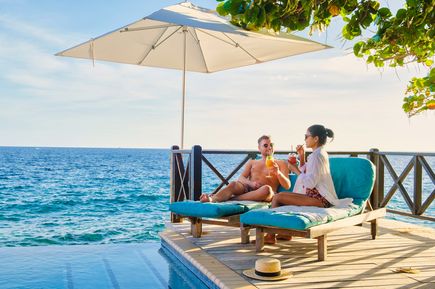 Zwei Personen sitzen auf einer Terrasse am Meer unter einem Sonnenschirm und trinken Cocktails neben einem Pool mit Blick auf den Ozean
