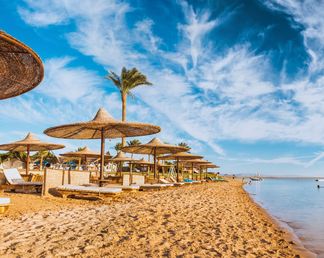 Strand am Roten Meer in Ägypten mit strohbedeckten Sonnenschirmen, Liegen und ruhigem, flach abfallendem Wasser unter blauem Himmel