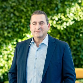 Porträt von Markus Förster im Außenbereich vor grünem Pflanzenhintergrund, stehend, gekleidet in Sakko und helles Hemd