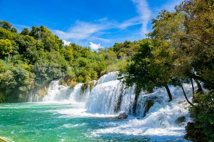 Kaskadenartige Wasserfälle im Nationalpark Krka in Kroatien, umgeben von dichtem Grün und türkisfarbenem Wasser unter blauem Himmel