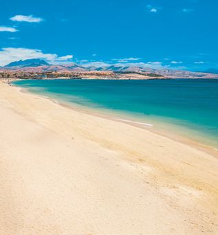 Breiter Sandstrand mit klarem, ruhigem Wasser und Bergen im Hintergrund unter blauem Himmel.