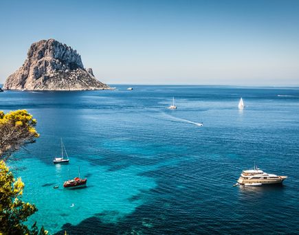 Blick auf das klare blaue Meer bei Ibiza mit mehreren Segelbooten und einer markanten Felseninsel im Hintergrund