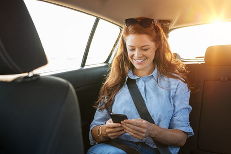 Frau mit blauer Bluse und Sonnenbrille auf dem Kopf sitzt angeschnallt auf dem Rücksitz eines Autos und hält ein Smartphone in den Händen