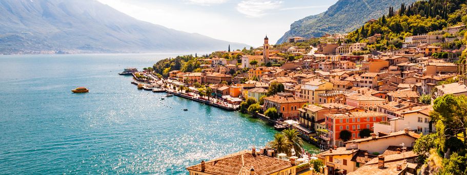 Blick auf eine belebte Uferstadt am Gardasee in Norditalien, umgeben von Bergen und klarem Wasser