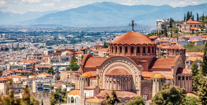 Blick auf die byzantinische Kirche mit roten Kuppeln in Thessaloniki, umgeben von Stadtgebäuden und Bergen im Hintergrund.
