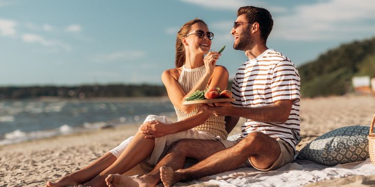 Paar sitzt bei einem Picknick am Strand, lacht gemeinsam und genießt frisches Obst mit Blick auf das Meer
