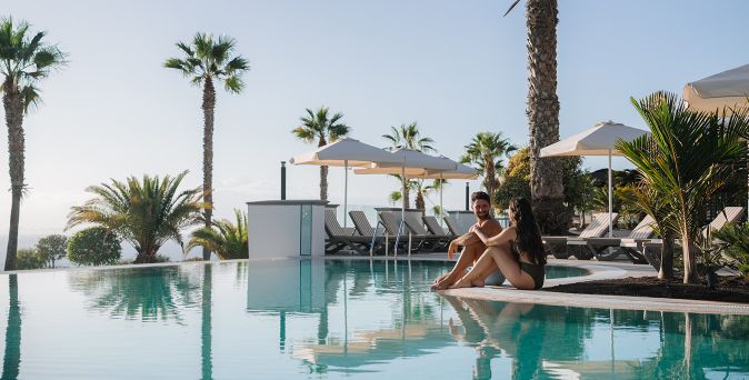 Zwei Personen sitzen am Rand eines Infinity-Pools mit Liegestühlen und Sonnenschirmen im Hintergrund, umgeben von Palmen.