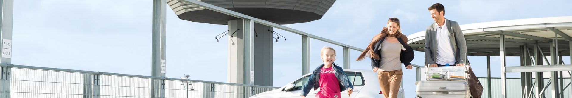 Familie mit Kind auf dem Parkplatz des Flughafens Hannover, unterwegs mit Rollkoffern und Gepäckwagen vor moderner Flughafenterminal-Architektur