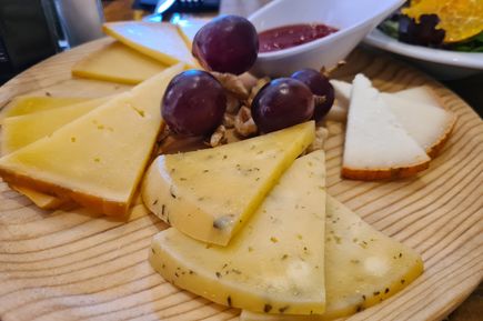 Auf einem Holzbrett liegen verschiedene Scheiben Queso de Mahón mit Kräutern und Naturrinde, dazu rote Trauben, Nüsse und eine Schale mit roter Soße