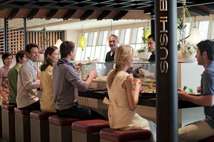 Gäste sitzen an der Sushi‑Bar der AIDAblu auf gepolsterten Hockern und beobachten die Zubereitung frischer Speisen hinter einer Glasvitrine