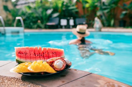 Frischer Obstkorb mit Wassermelone, Mango und Mangostan am Poolrand, im Hintergrund Person im Pool