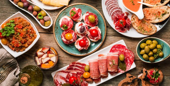 Bunte Auswahl spanischer Tapas auf Holztisch: Brot mit Tomate, Wurst- und Schinkenscheiben, Oliven, Ratatouille, Käse, Sangria und kleine belegte Häppchen mit Tomaten