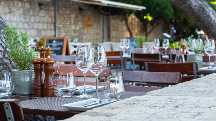 Stilvoll gedeckte Tische im Außenbereich eines Restaurants mit Weingläsern, Besteck und Kräutern auf Holztischen