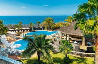Hotelpool mit Liegestühlen und Sonnenschirmen, umgeben von Palmen und einer Strohdachbar, im Hintergrund das Meer unter blauem Himmel.