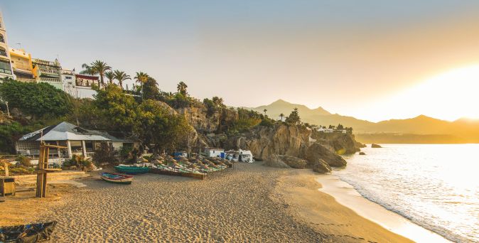 Sonnenuntergang über einem Sandstrand in Andalusien mit Felsen, kleinen Booten und Häusern am Hang