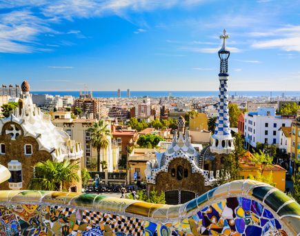 Aussicht über den Park Güell in Barcelona mit den farbenfrohen Mosaiken von Antoni Gaudí, den markanten Gebäuden am Parkeingang und dem Panorama der Stadt bis hin zum Mittelmeer