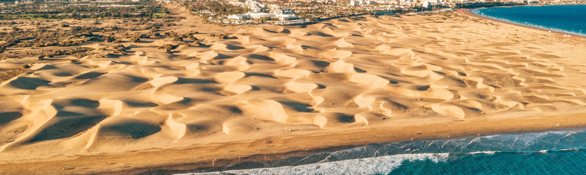 Luftaufnahme von Sanddünen an der Küste mit dahinter liegender Stadt und Bergen im Hintergrund