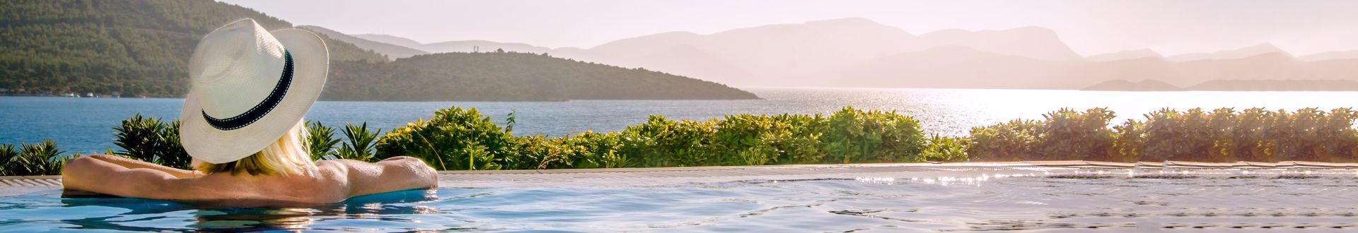 Person entspannt im Infinity-Pool mit Blick auf das Meer und grüne Hügel, sonniger Urlaubstag in mediterraner Landschaft
