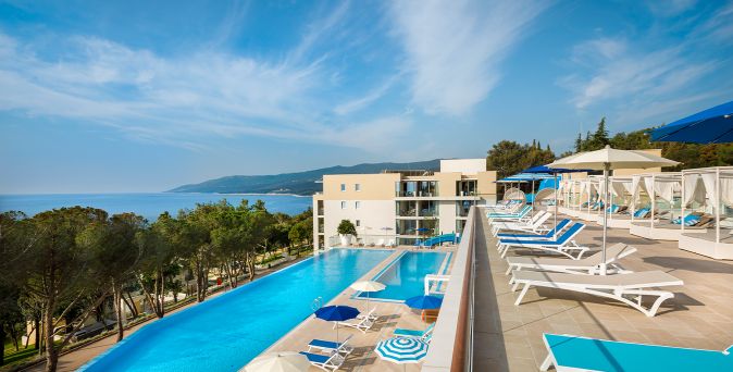 Infinity-Pool mit Sonnenterrasse und Liegen im Girandella Maro Suites Resort, umgeben von Pinienwald mit Blick auf die Adria.