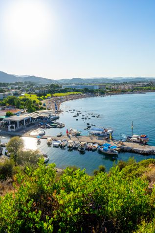 Blick auf den Hafen von Afántou auf Rhodos mit mehreren Booten am Steg, Uferpromenade und dahinter liegenden Gebäuden vor Bergkulisse.