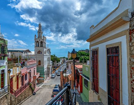 Blick auf eine belebte Straße mit bunten Häusern und einer weißen Kirche mit Turm unter einem bewölkten Himmel.