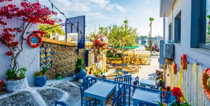 Blick auf eine sonnige Außenterrasse eines Restaurants mit blauen Stühlen, weißen Tischen, blühenden roten Bougainvillea und Blick auf den Hafen.