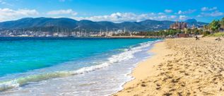 Sandstrand an der Playa de Palma auf Mallorca mit türkisblauem Meer, sanften Wellen, Palmenpromenade, Yachthafen und Bergkulisse im Hintergrund