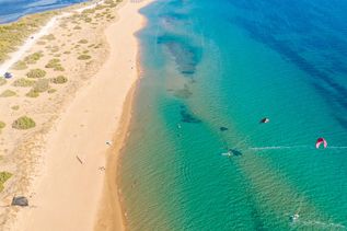 Luftaufnahme des Issos-Strands auf Korfu mit Sanddünen links und klarem, türkisfarbenem Wasser rechts, in dem mehrere Kitesurfer aktiv sind.