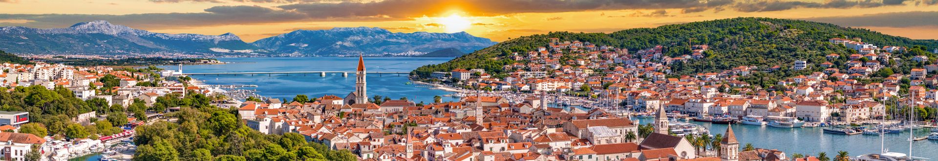 Panorama der historischen Altstadt von Trogir in Kroatien mit roten Ziegeldächern, Kirchtürmen, Yachthafen, Meeresbucht und Bergen im Hintergrund bei Sonnenuntergang