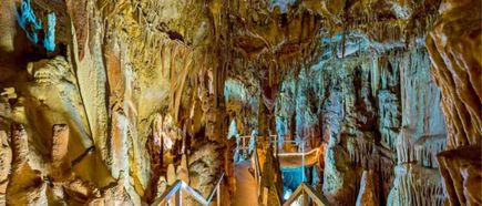 Beleuchteter Holzsteg mit Geländer führt durch eine Höhle mit zahlreichen Stalaktiten und Stalagmiten an den Wänden und der Decke.