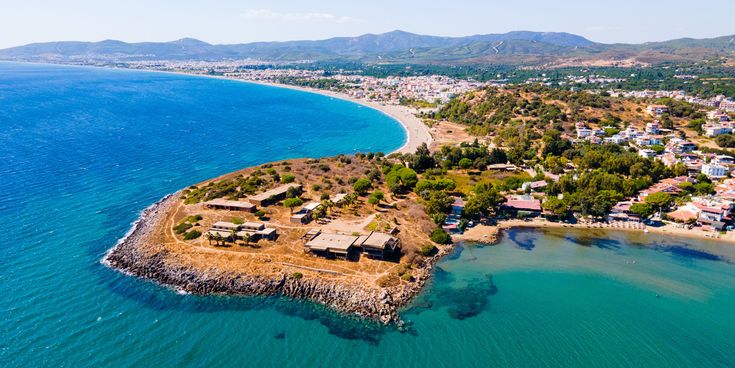 Luftaufnahme einer türkisblauen Bucht bei Izmir in der Türkei: lange Sandstrand-Kurve, kleine Landzunge mit Gebäuden, Küstenort und grüne Berge im Hintergrund