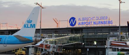 Ein KLM-Flugzeug steht auf dem Vorfeld des Albrecht Dürer Airport Nürnberg vor dem Terminalgebäude mit der sichtbar beleuchteten Aufschrift „ALBRECHT DÜRER AIRPORT NÜRNBERG“. Geben Sie Ihr Feedback zu BizChat ab
