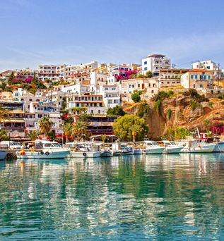 Blick auf eine Küstenstadt mit weißen Gebäuden und bunten Blumen, umgeben von Wasser und Booten im Vordergrund.