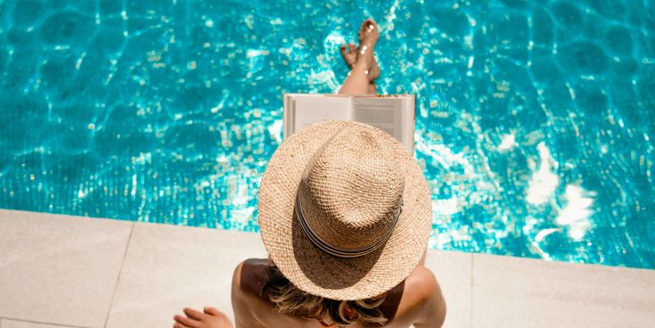 Frau mit Strohhut sitzt am Pool und liest ein Buch, während ihre Füße im klaren, türkisblauen Wasser hängen