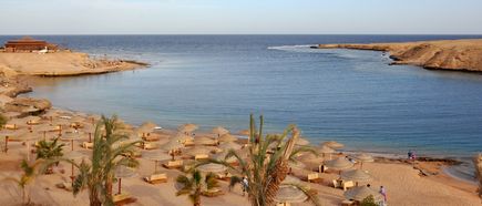 Sandstrand in der Makadi Bay in Ägypten mit Palmen, zahlreichen Sonnenschirmen und Liegen vor dem ruhigen blauen Meer, umgeben von felsigen Küsten