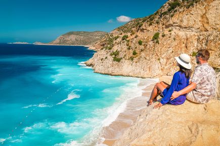 Paar sitzt auf einem Felsen hoch über einem türkisfarbenen Strand und blickt auf das Meer und die steilen Klippen